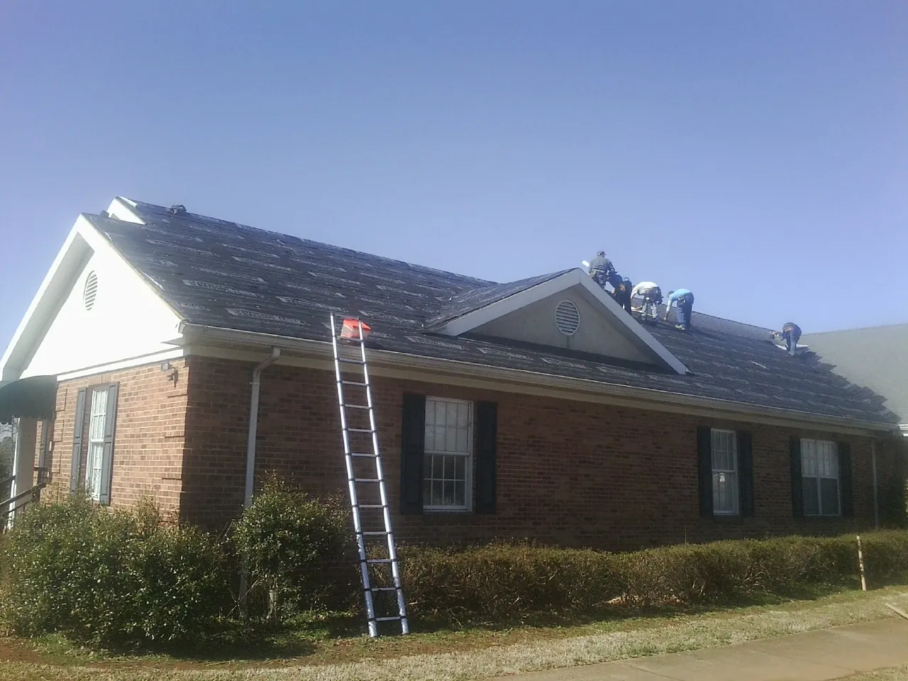 Crew tearing shingles off a brick ranch roof with a ladder set — Metal Roof Repair work by our crew in Rock Rapids