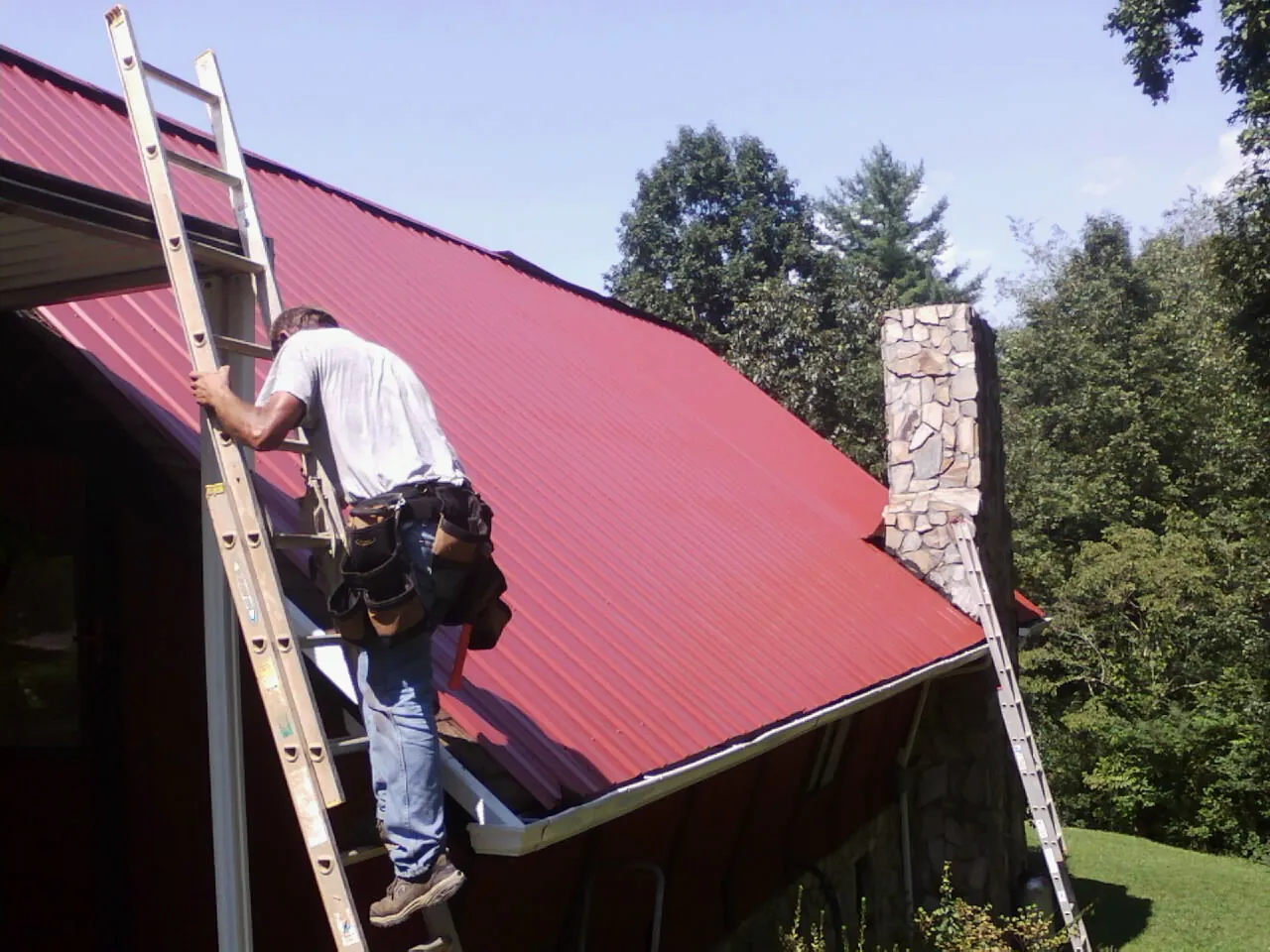 Roofer on a ladder beside a red metal roof and stone chimney — Soffit & Fascia Repair by Roofing Brothers in Rock Rapids